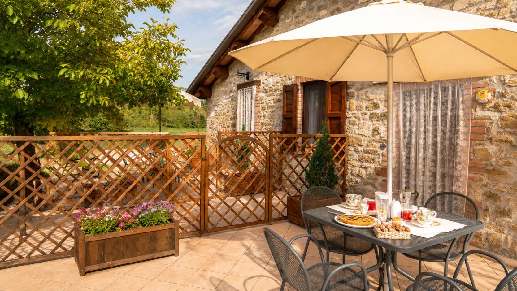 Terrazza del''appartamento Girasole ina agriturismo umbro Domus Santa Croce. Colazione all'aperto con ombrelloni, tavolo e sedie in ferro, immersa nel verde e fiori.